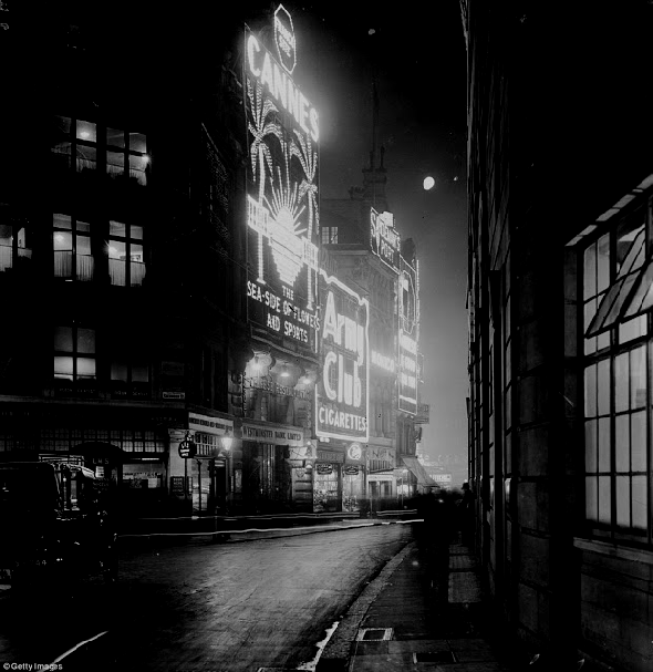 Black and white picture of 1920s city street at night with large illuminated adverts on the sides of several of the buildings.